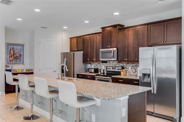 kitchen featuring appliances with stainless steel finishes, a kitchen breakfast bar, light tile patterned floors, light stone counters, and a center island with sink