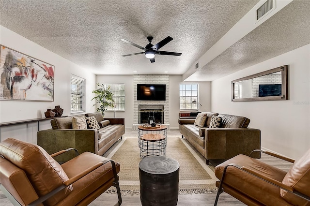 living room with light wood-type flooring, ceiling fan, a brick fireplace, and a textured ceiling