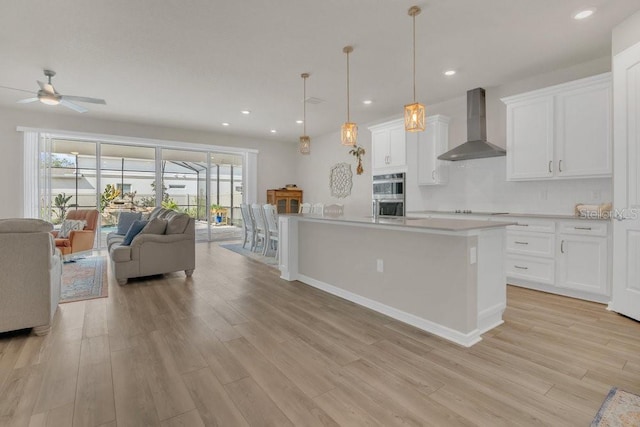 kitchen featuring pendant lighting, white cabinetry, wall chimney range hood, a center island with sink, and light wood-type flooring