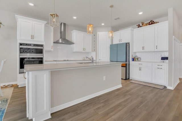 kitchen featuring an island with sink, sink, white cabinets, hanging light fixtures, and wall chimney range hood