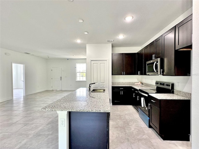 kitchen with sink, appliances with stainless steel finishes, dark brown cabinetry, light stone countertops, and a center island with sink