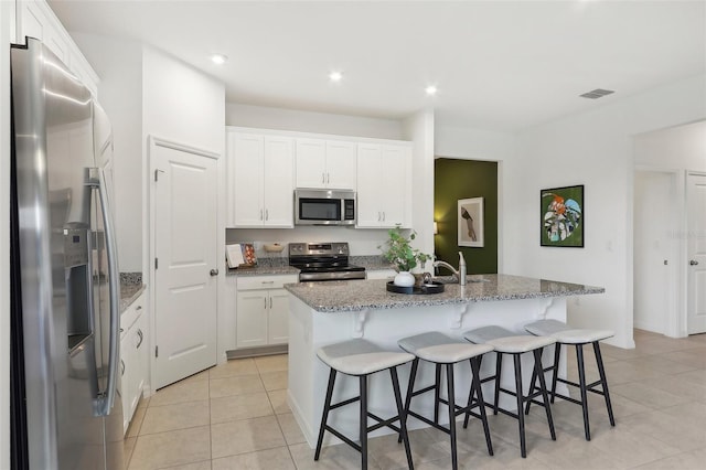 kitchen featuring light stone countertops, white cabinets, appliances with stainless steel finishes, a kitchen island with sink, and light tile patterned floors