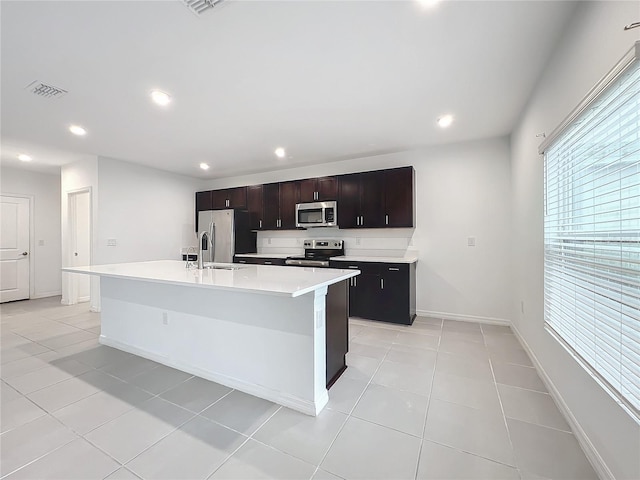 kitchen with stainless steel appliances, dark brown cabinetry, a center island with sink, and light tile patterned flooring