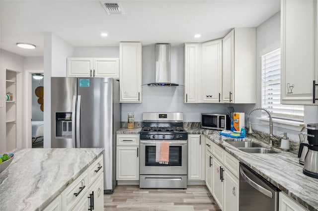 kitchen featuring light stone countertops, appliances with stainless steel finishes, white cabinets, wall chimney range hood, and sink