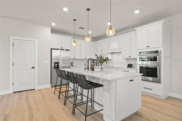 kitchen featuring decorative light fixtures, white cabinetry, stainless steel appliances, a kitchen island with sink, and custom range hood