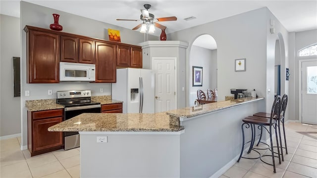 kitchen with light tile patterned flooring, a kitchen bar, ceiling fan, white appliances, and light stone countertops