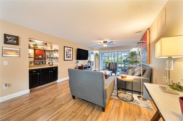 living room with ceiling fan, a textured ceiling, indoor wet bar, and light wood-type flooring