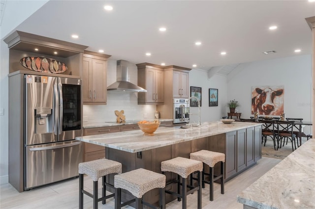 kitchen with appliances with stainless steel finishes, a large island, light stone countertops, decorative backsplash, and wall chimney range hood