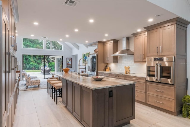 kitchen with a breakfast bar, appliances with stainless steel finishes, light stone countertops, an island with sink, and wall chimney exhaust hood