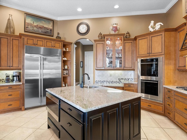 kitchen with light tile patterned flooring, sink, built in appliances, a center island with sink, and light stone countertops