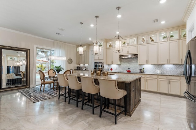 kitchen featuring black refrigerator, pendant lighting, an island with sink, light stone counters, and cream cabinetry