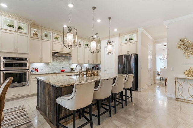 kitchen with sink, an island with sink, stainless steel appliances, light stone countertops, and backsplash