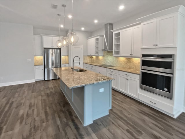 kitchen with wall chimney range hood, sink, stainless steel appliances, white cabinets, and a center island with sink