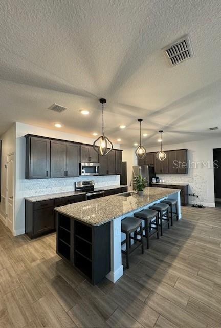 kitchen featuring backsplash, hanging light fixtures, a kitchen island with sink, dark brown cabinetry, and stainless steel appliances