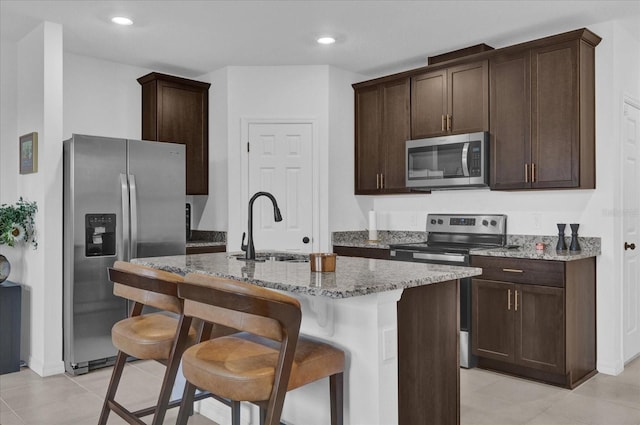 kitchen featuring light stone counters, stainless steel appliances, dark brown cabinets, an island with sink, and a kitchen bar