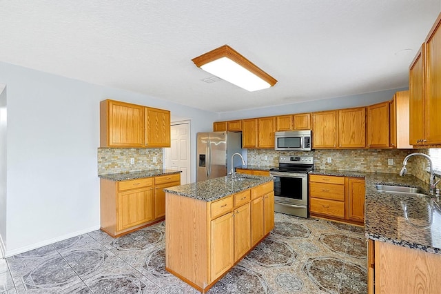 kitchen with sink, appliances with stainless steel finishes, an island with sink, decorative backsplash, and dark stone counters