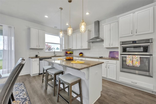 kitchen with wall chimney range hood, white cabinetry, stainless steel appliances, a center island, and stone countertops