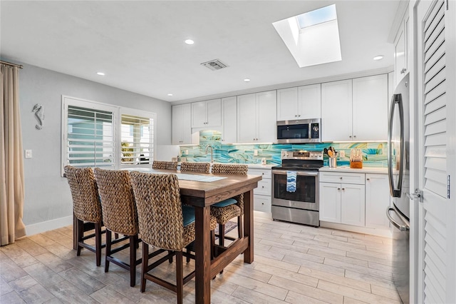 kitchen with a skylight, white cabinets, decorative backsplash, stainless steel appliances, and light hardwood / wood-style flooring