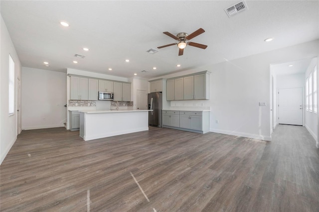 kitchen with dark wood-type flooring, gray cabinets, stainless steel appliances, an island with sink, and decorative backsplash