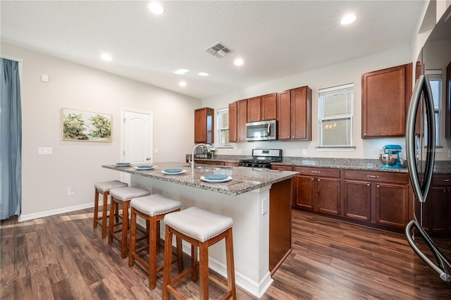 kitchen with appliances with stainless steel finishes, dark hardwood / wood-style floors, a breakfast bar, a kitchen island with sink, and light stone countertops