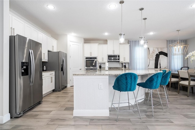 kitchen with appliances with stainless steel finishes, decorative light fixtures, white cabinets, light stone counters, and a center island with sink