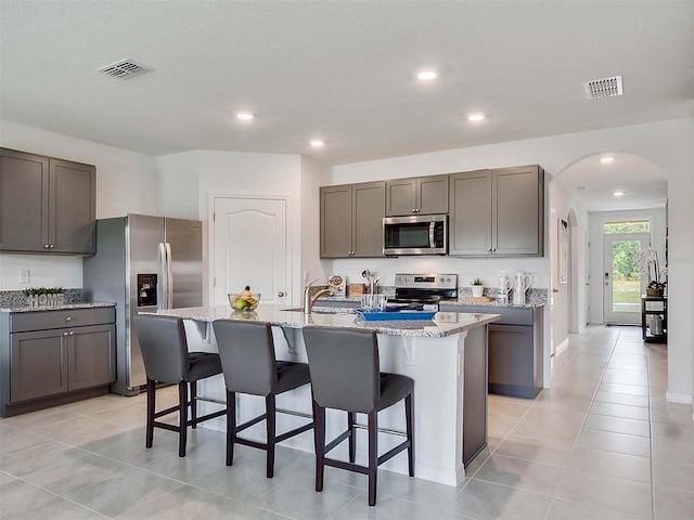 kitchen featuring a breakfast bar area, light tile patterned floors, an island with sink, stainless steel appliances, and light stone countertops