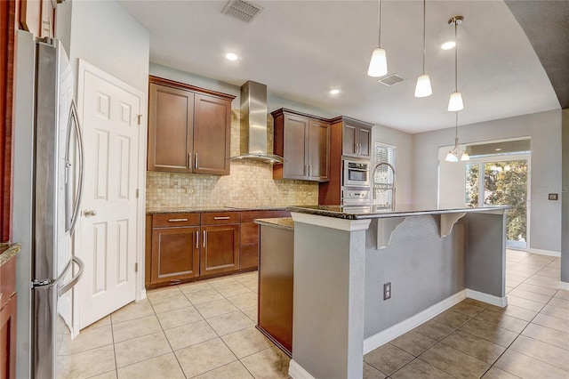 kitchen with an island with sink, stainless steel appliances, dark stone counters, wall chimney exhaust hood, and pendant lighting