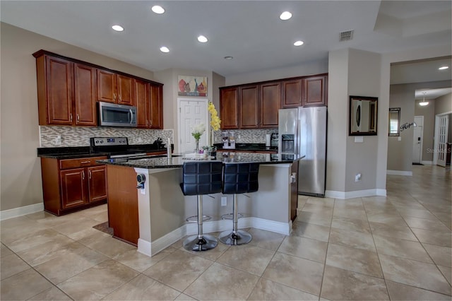 kitchen with light tile patterned floors, dark stone countertops, stainless steel appliances, an island with sink, and a kitchen bar