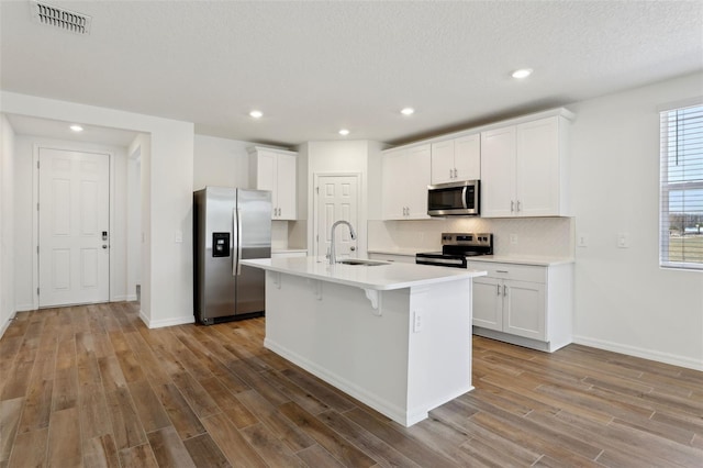 kitchen with appliances with stainless steel finishes, white cabinetry, wood-type flooring, sink, and a kitchen island with sink