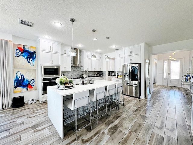 kitchen featuring a breakfast bar, hanging light fixtures, stainless steel appliances, an island with sink, and wall chimney exhaust hood