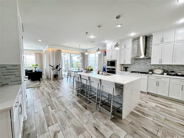 kitchen with white cabinetry, hanging light fixtures, a center island with sink, a kitchen breakfast bar, and wall chimney range hood