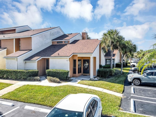 view of property with a front lawn, stucco siding, uncovered parking, and a shingled roof