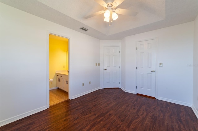 unfurnished bedroom with a tray ceiling, visible vents, ensuite bathroom, dark wood-type flooring, and baseboards