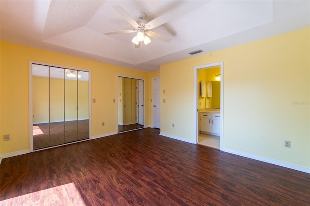 unfurnished bedroom with a tray ceiling, dark wood-style flooring, multiple closets, visible vents, and baseboards