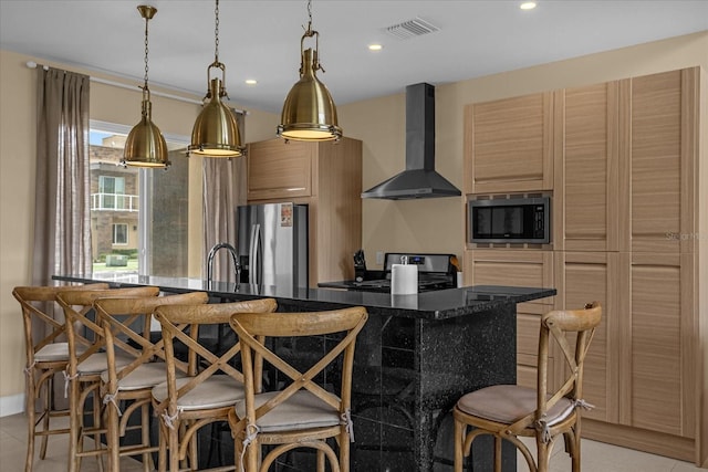 kitchen with wall chimney range hood, a breakfast bar, appliances with stainless steel finishes, a kitchen island with sink, and light brown cabinetry