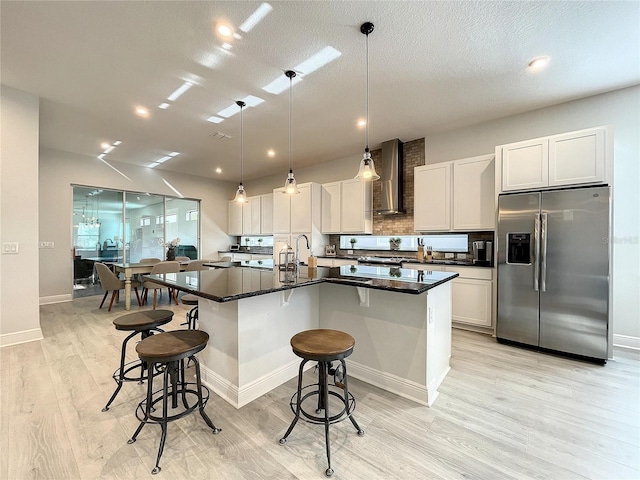 kitchen featuring stainless steel refrigerator with ice dispenser, a breakfast bar area, white cabinetry, a kitchen island with sink, and wall chimney range hood