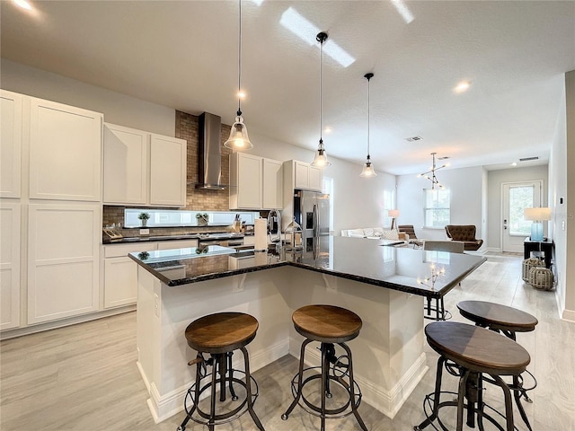 kitchen featuring pendant lighting, wall chimney range hood, a kitchen island with sink, white cabinetry, and a kitchen bar