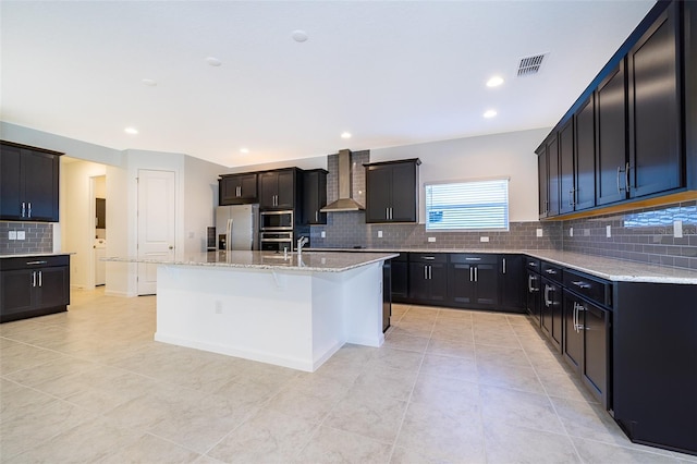 kitchen featuring wall chimney range hood, light tile patterned floors, appliances with stainless steel finishes, a kitchen island with sink, and light stone countertops