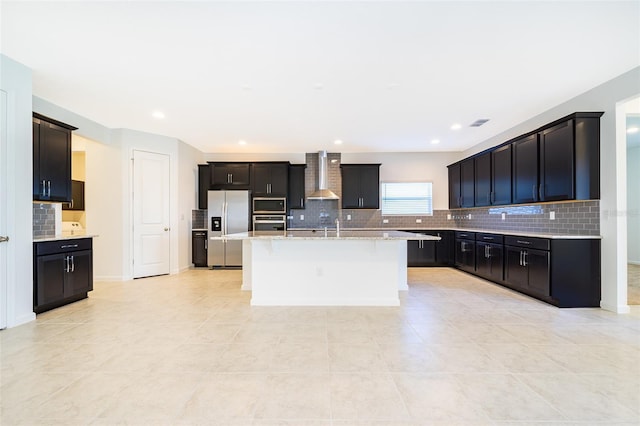 kitchen with an island with sink, sink, light tile patterned floors, stainless steel appliances, and wall chimney range hood