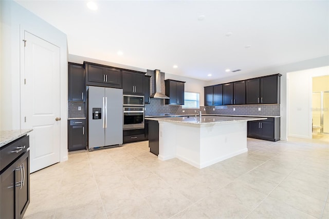 kitchen featuring light stone counters, built in appliances, a kitchen island with sink, and wall chimney range hood