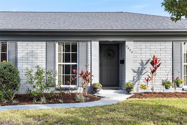 view of exterior entry featuring a yard, brick siding, and roof with shingles