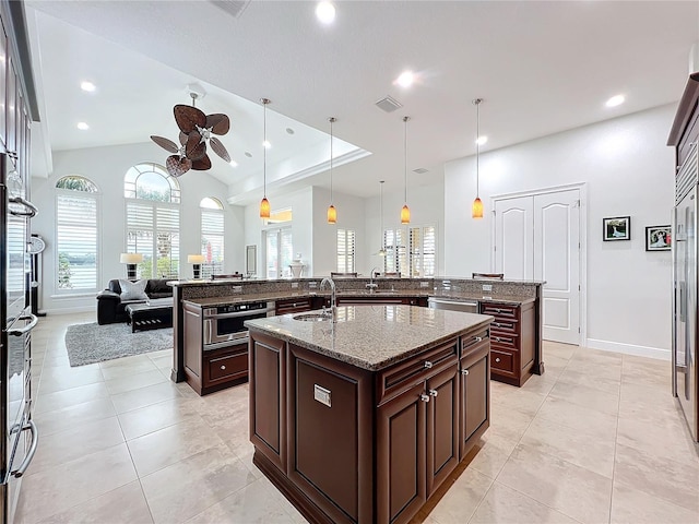 kitchen with pendant lighting, sink, dark brown cabinets, light stone counters, and an island with sink