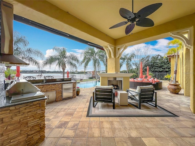 view of patio with an outdoor living space with a fireplace, sink, exterior kitchen, ceiling fan, and a water view