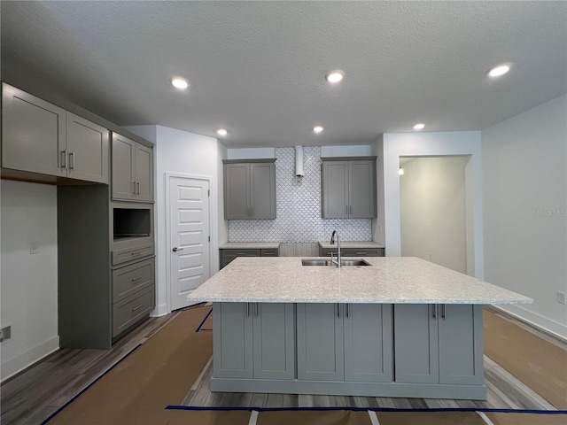 kitchen featuring gray cabinetry, a kitchen island with sink, a sink, decorative backsplash, and dark wood-style flooring