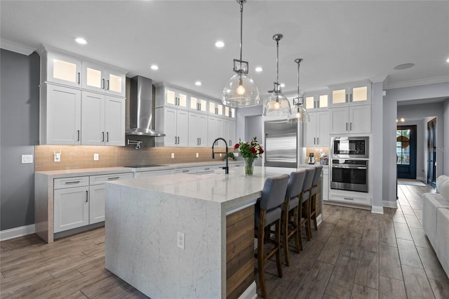 kitchen featuring wall chimney exhaust hood, built in appliances, a center island with sink, and white cabinets