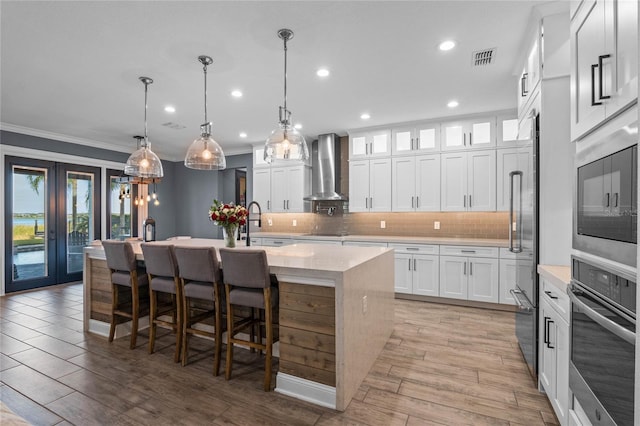 kitchen featuring decorative light fixtures, white cabinetry, an island with sink, stainless steel appliances, and wall chimney exhaust hood