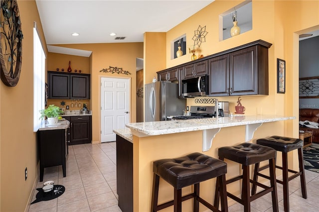 kitchen with dark brown cabinetry, a breakfast bar, vaulted ceiling, light tile patterned floors, and appliances with stainless steel finishes