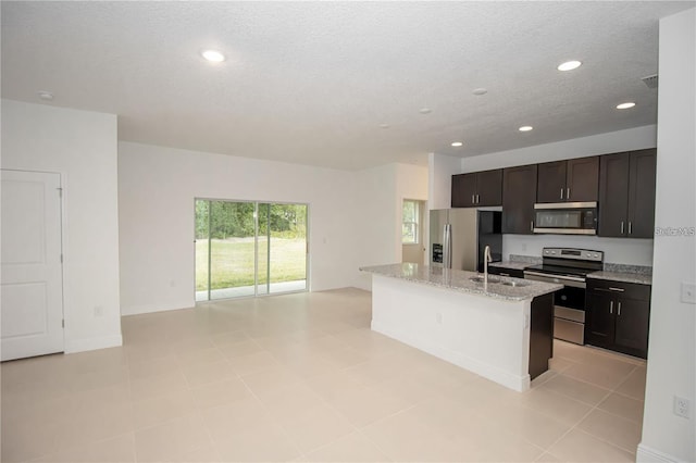 kitchen featuring sink, appliances with stainless steel finishes, light stone counters, dark brown cabinetry, and an island with sink