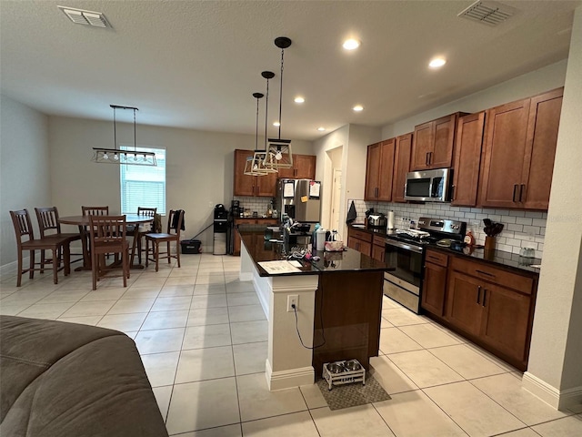 kitchen featuring light tile patterned flooring, an island with sink, backsplash, hanging light fixtures, and stainless steel appliances