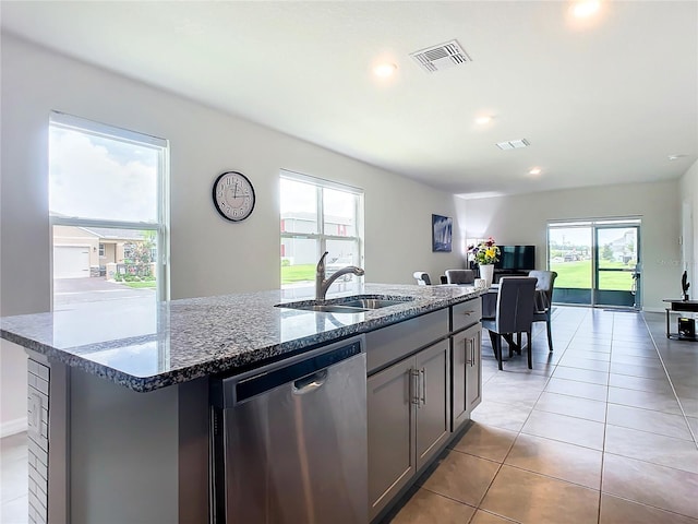 kitchen with sink, light tile patterned floors, stainless steel dishwasher, dark stone counters, and a kitchen island with sink